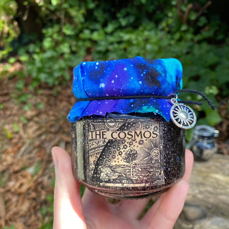 May include: A black glass jar with a galaxy print fabric lid and a silver charm. The jar has a label that reads "The Cosmos".