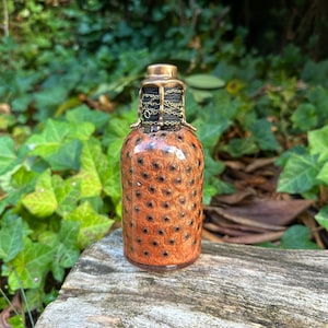 May include: A small, brown glass bottle with a gold metal top and a black chain. The bottle has a speckled pattern.