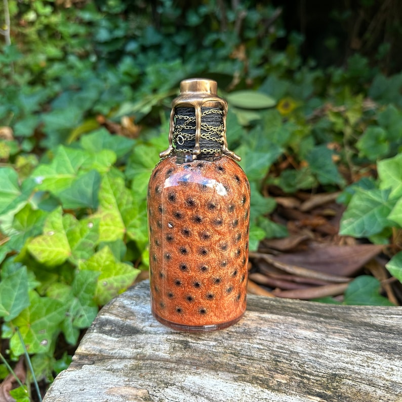 May include: A small, brown glass bottle with a gold metal top and a black chain. The bottle has a speckled pattern.