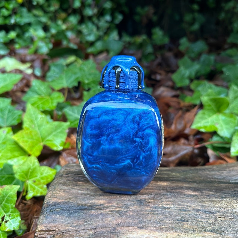 May include: A small, square, blue glass bottle with a black, dripping, resin lid. The bottle has a metallic, iridescent finish.
