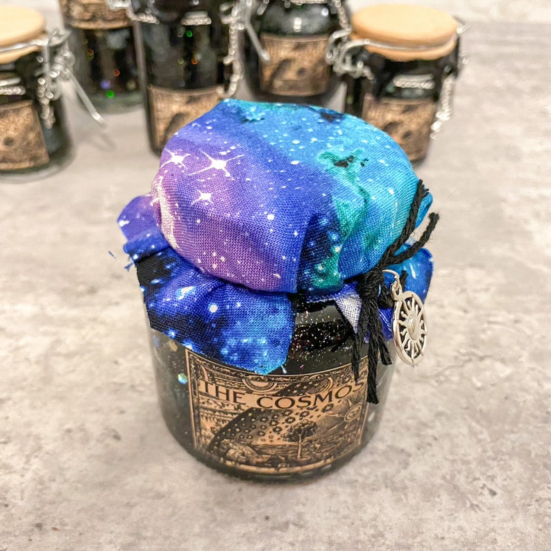 May include: A small glass jar with a black lid and a galaxy-themed fabric cover. The jar has a label that reads "The Cosmos".