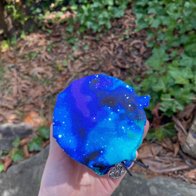 May include: A small round container with a blue, green, and black galaxy print fabric cover. A silver charm with a sun design is attached to the fabric cover.