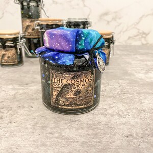May include: A small glass jar with a black and silver lid, filled with black glitter and a galaxy-themed fabric tied around the top. The jar has a label that reads "THE COSMOS" with a vintage-style illustration of a celestial scene.