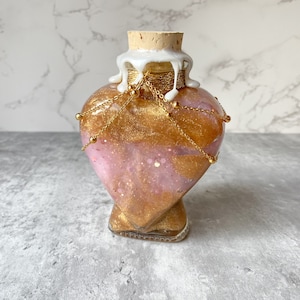 May include: A heart-shaped glass bottle with a gold glitter and pink swirl design. The bottle has a cork stopper and is decorated with gold chains and beads.