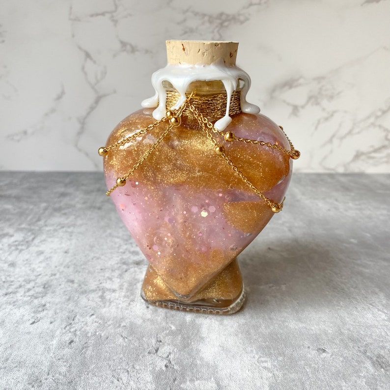 May include: A heart-shaped glass bottle with a gold glitter and pink swirl design. The bottle has a cork stopper and is decorated with gold chains and beads.