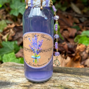 K&ouml;nnte beinhalten: Eine kleine Glasflasche mit einer violetten Fl&uuml;ssigkeit und einem Etikett mit der Aufschrift "Simple. Yet Powerful. SLEEPING DRAUGHT.  Instantaneous Sleep." Die Flasche hat eine goldene Kette mit violetten Perlen und einen goldenen Anh&auml;nger.