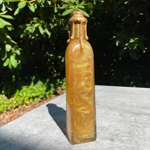 K&ouml;nnte beinhalten: Eine klare Glasflasche, gef&uuml;llt mit schimmernder goldener Fl&uuml;ssigkeit. Die Flasche hat einen goldfarbenen, strukturierten Verschluss mit Wachstropfeneffekt. Die Flasche ist rechteckig mit abgerundeten Kanten, vor verschwommenem gr&uuml;nem Laub.