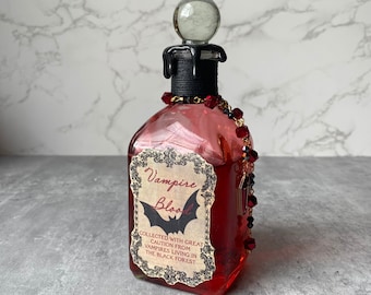 May include: A glass bottle with a black stopper and a red liquid inside. The bottle has a label that reads "Vampire Blood" with a black bat illustration. The label also says "Collected with great caution from vampires living in the Black Forest."