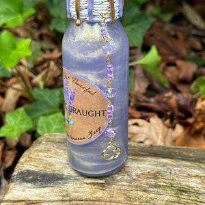 K&ouml;nnte beinhalten: Eine kleine Glasflasche mit einem lila und goldenen Etikett, auf dem "Yet Powerful Draught for Spontaneous Sleep" steht. Die Flasche hat einen goldenen Anh&auml;nger und ist mit einer glitzernden Fl&uuml;ssigkeit gef&uuml;llt.