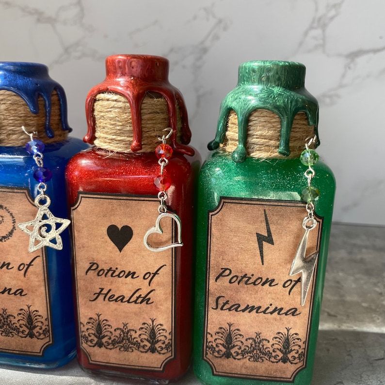 May include: Three glass potion bottles with cork stoppers and labels. The bottles are filled with glitter and have a red, green, and blue liquid. Each bottle has a charm hanging from it. The labels read "Potion of Health", "Potion of Stamina", and "Potion of Strength".