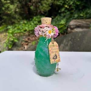 May include: A green glass bottle with a cork stopper, decorated with pink and white flowers. A tag reading "Flora's Garden" is attached, with a bee charm. The bottle has a textured surface. The bottle is approximately 15 cm tall.