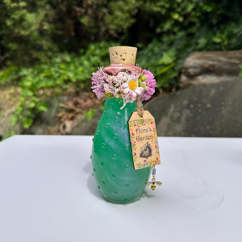 May include: A green glass bottle with a cork stopper, decorated with pink and white flowers. A tag reading "Flora's Garden" is attached, with a bee charm. The bottle has a textured surface. The bottle is approximately 15 cm tall.