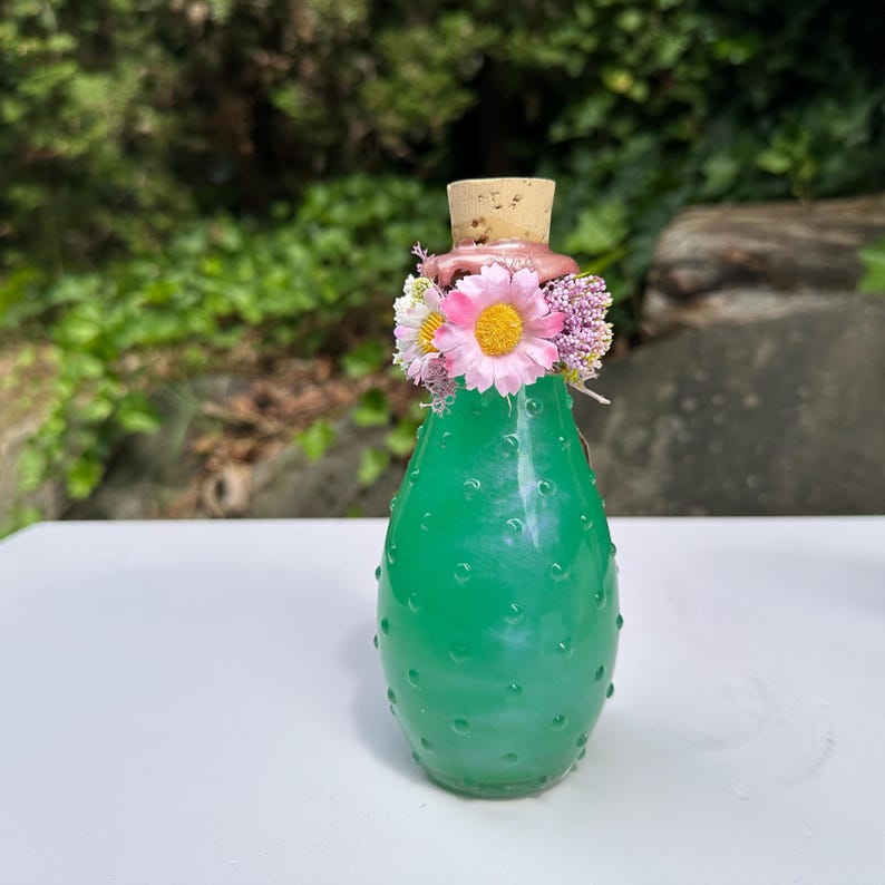 May include: A green glass bottle with a cactus design, featuring a cork stopper and a floral arrangement of pink, white, and purple flowers. The bottle sits on a white surface with a blurred green backdrop.