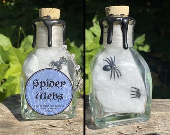 Puede incluir: Una pequeña botella de vidrio con un tapón de corcho y cera negra goteando por los lados. La botella está llena de algodón blanco y contiene dos arañas de plástico negro. Una etiqueta en la parte delantera de la botella dice "Telarañas Se pueden usar para atar y atrapar hechizos."