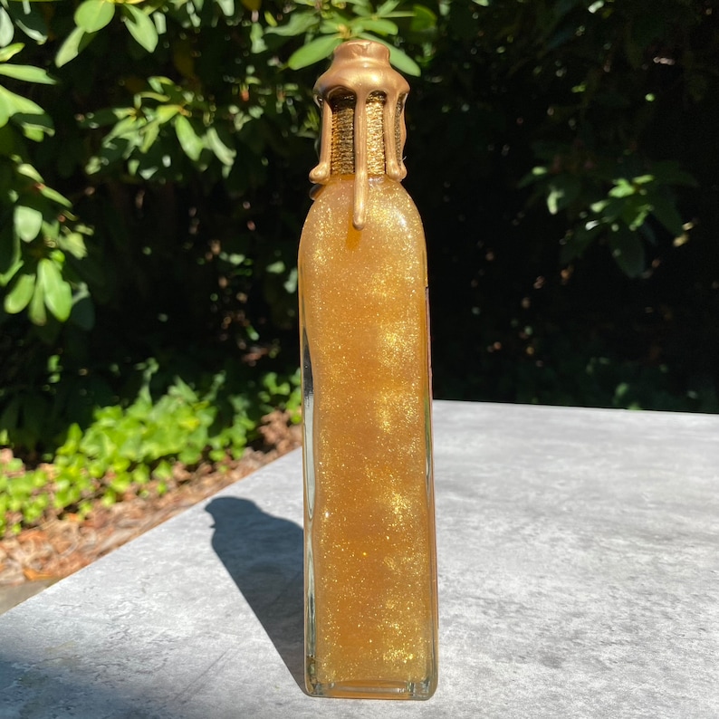 K&ouml;nnte beinhalten: Eine hohe, rechteckige Glasflasche, gef&uuml;llt mit glitzernder, goldfarbener Fl&uuml;ssigkeit. Die Flasche hat einen dekorativen, goldfarbenen Verschluss. Die Flasche steht auf einer grauen Oberfl&auml;che mit gr&uuml;nem Hintergrund.