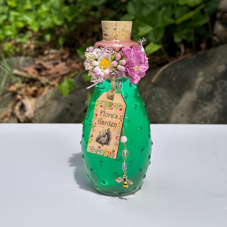 May include: A green glass bottle with a cork stopper, decorated with pink and white flowers. A tag reading "Flora's Garden" is attached, along with a beaded charm and a bee pendant. The bottle has a textured surface and is likely for decorative purposes.