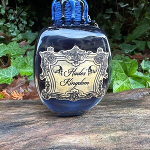 May include: A black glass bottle with a blue and gold label that reads "Hades' Kingdom". The bottle has a skull design on the label and is dripping with a black liquid.