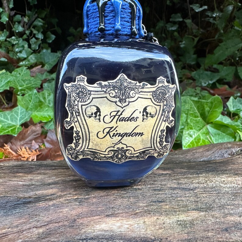 May include: A black glass bottle with a blue and gold label that reads "Hades' Kingdom". The bottle has a skull design on the label and is dripping with a black liquid.