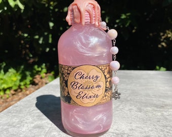 May include: A pink glass bottle with a pink and gold label that reads "Cherry Blossom Elixir". The bottle has a pink wax seal and a silver chain with white beads hanging from the top.