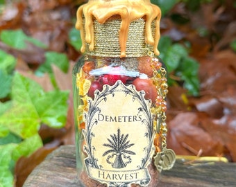 Puede incluir: Un frasco de vidrio con una tapa dorada y una etiqueta que dice "Demeter's Harvest". El frasco está lleno de varias hierbas secas, bayas y otros ingredientes naturales.