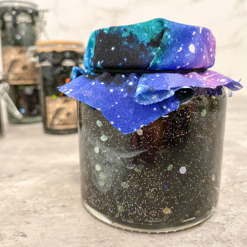 May include: A glass jar filled with black glitter and a blue and purple galaxy print fabric tied around the lid.