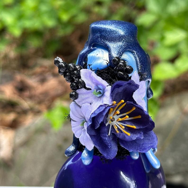 May include: A dark blue decorative bottle with a metallic finish, decorated with purple artificial flowers and black berries. The bottle has a dripped wax effect on the top. The bottle is approximately 15 cm tall.