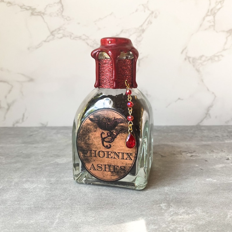 Puede incluir: Una botella de vidrio con un sello de cera roja y un amuleto rojo y dorado. La botella est&aacute; llena de polvo negro y tiene una etiqueta que dice "PHOENIX ASHES".