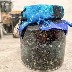 May include: A glass jar with a black glittery liquid inside, topped with a blue and white fabric with a star pattern. The jar has a silver charm attached to the fabric.