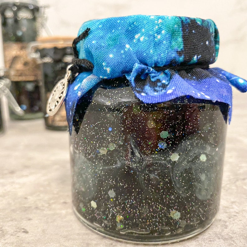 May include: A glass jar with a black glittery liquid inside, topped with a blue and white fabric with a star pattern. The jar has a silver charm attached to the fabric.