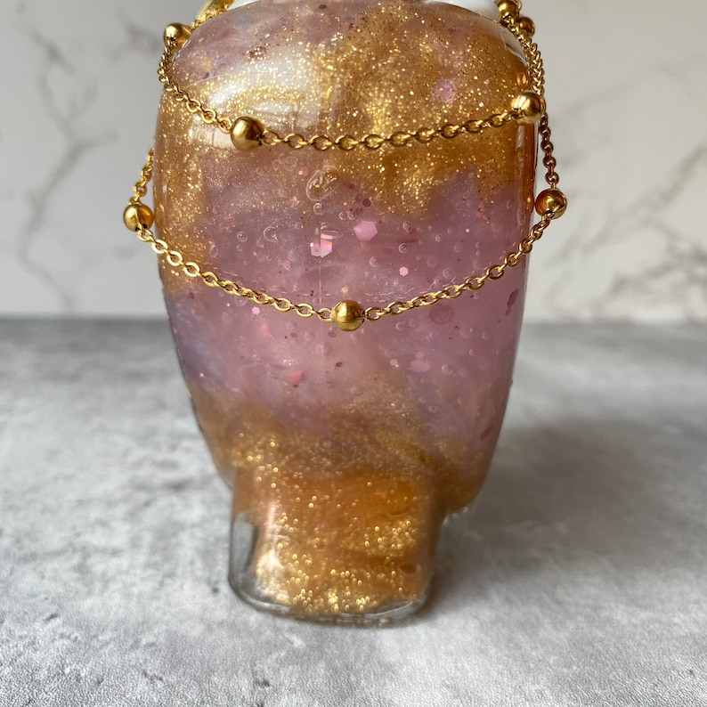 May include: A glass bottle with a pink and gold glitter design. The bottle is decorated with two gold chains with small gold beads.