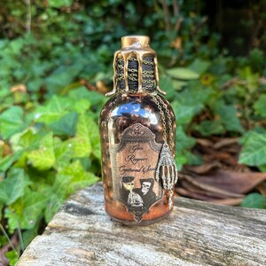 May include: A small glass bottle with a gold-colored metal lid and a label that reads "Grim Reaper's Captured Soul". The bottle is decorated with a skeleton holding a glass of liquid and a dangling skeleton hand charm.