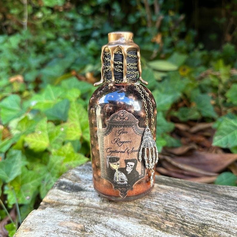 May include: A small glass bottle with a gold-colored metal lid and a label that reads "Grim Reaper's Captured Soul". The bottle is decorated with a skeleton holding a glass of liquid and a dangling skeleton hand charm.