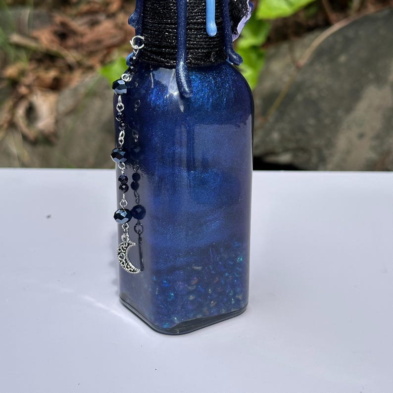 May include: A dark blue, square bottle with a shimmering, glittery interior. The bottle has a black cord around the neck and a beaded charm with a crescent moon pendant. The bottle is filled with small, dark blue particles.