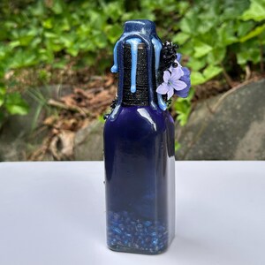 May include: A dark blue glass bottle with a decorative top. The top is wrapped in black twine and adorned with blue wax drips and small purple flowers. The bottle is filled with blue beads.
