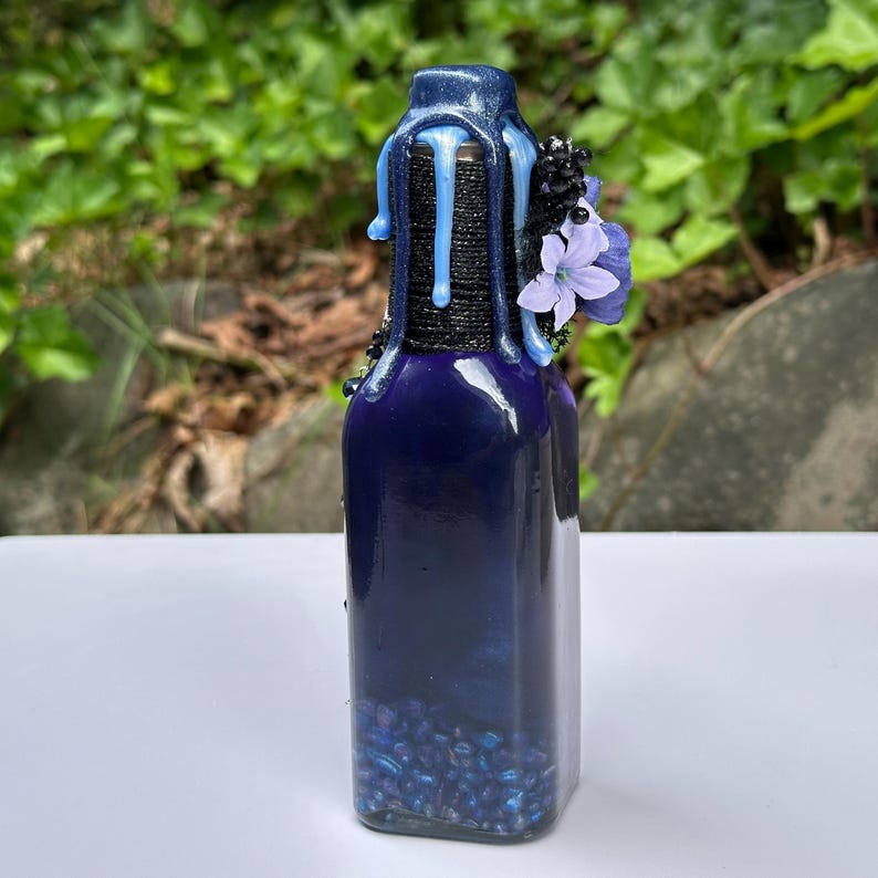 May include: A dark blue glass bottle with a decorative top. The top is wrapped in black twine and adorned with blue wax drips and small purple flowers. The bottle is filled with blue beads.