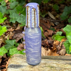 K&ouml;nnte beinhalten: Eine kleine Glasflasche mit einem schimmernden Finish in Lila und Gold. Die Flasche hat einen gold- und wei&szlig; gestreiften Verschluss mit einem violetten Tropfeffekt. Eine lila und goldene Kette h&auml;ngt an der Flasche.