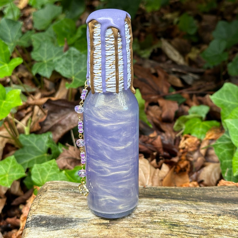 K&ouml;nnte beinhalten: Eine kleine Glasflasche mit einem schimmernden Finish in Lila und Gold. Die Flasche hat einen gold- und wei&szlig; gestreiften Verschluss mit einem violetten Tropfeffekt. Eine lila und goldene Kette h&auml;ngt an der Flasche.