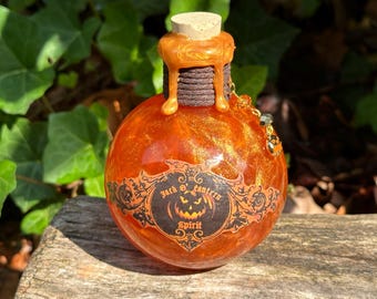 Può includere: Una piccola bottiglia rotonda di colore arancione con glitter dorati e un tappo di sughero. La bottiglia ha un'etichetta che dice "Jack O' Lantern Spirit".