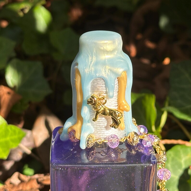 May include: A blue and gold bottle stopper with a gold lion charm. The bottle stopper is decorated with a white string and purple beads.