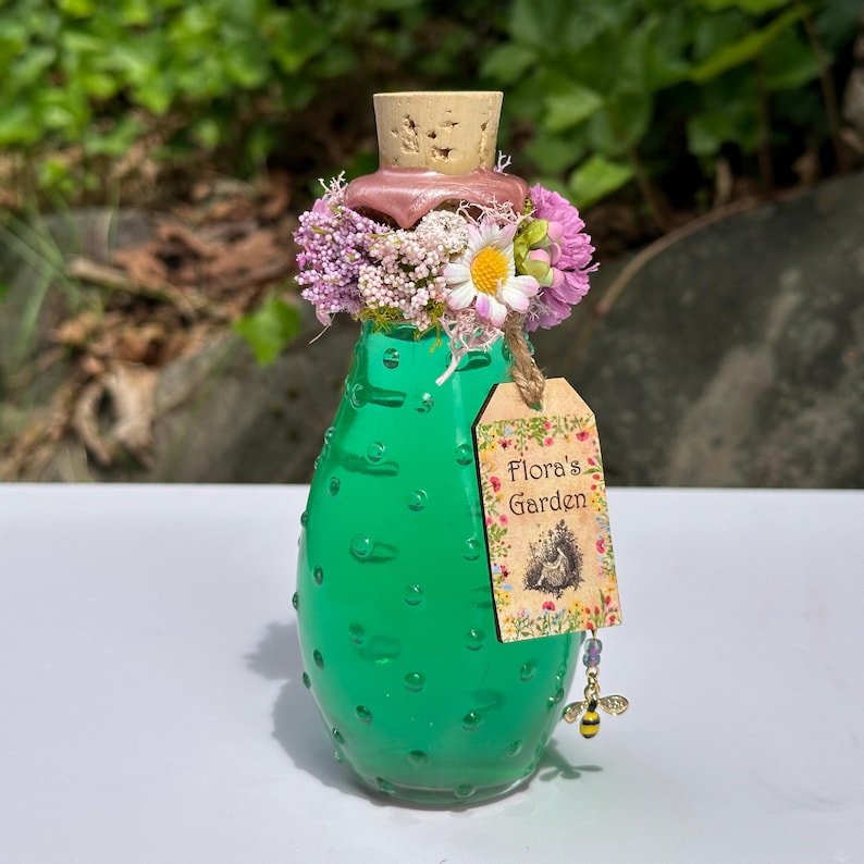 May include: A green glass bottle with a cork stopper, decorated with a floral arrangement of pink and white flowers. A tag reading "Flora's Garden" hangs from the bottle, with a bee charm.