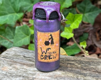 May include: A purple glitter bottle with a label that reads "Witches Brew". The label features a witch stirring a cauldron. The bottle has a black rope around the neck and a purple wax seal with a small silver bell.