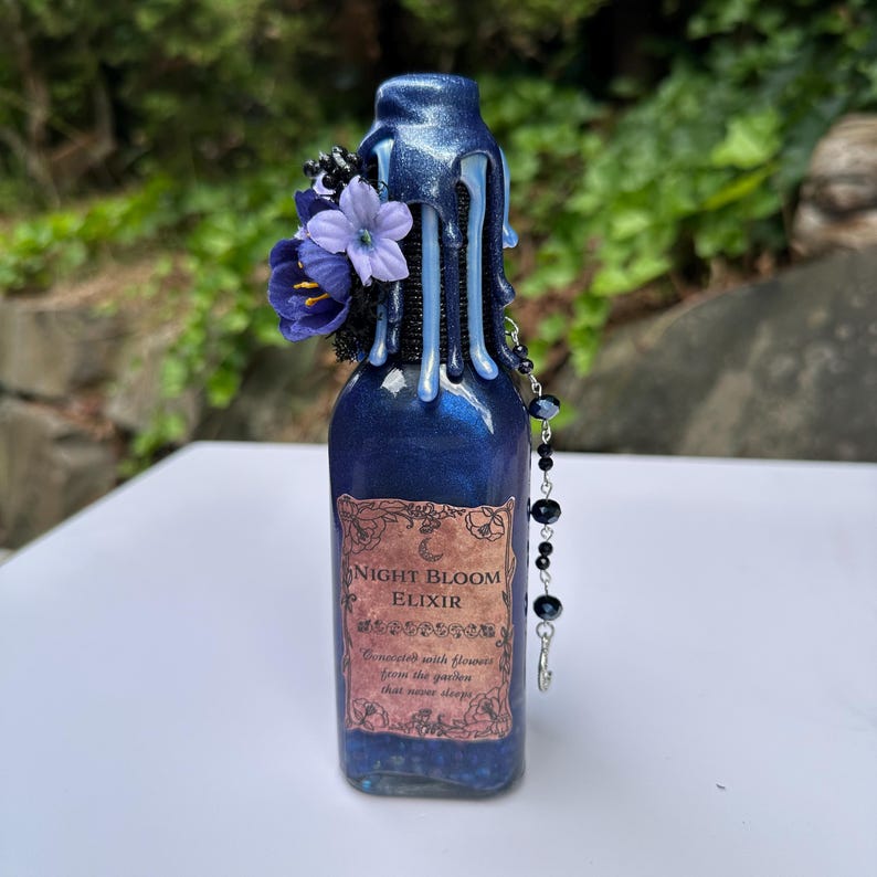May include: A dark blue glass bottle with a dripped wax top and a floral arrangement. The label reads "Night Bloom Elixir". A beaded charm hangs from the bottle. The bottle is approximately 15 cm tall.
