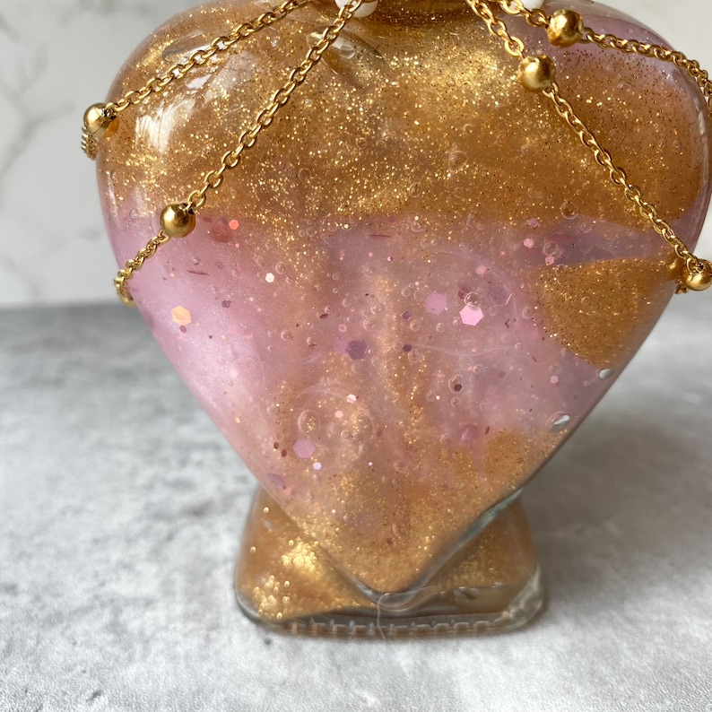 May include: A heart-shaped glass bottle filled with pink and gold glitter, adorned with a gold chain and beads.