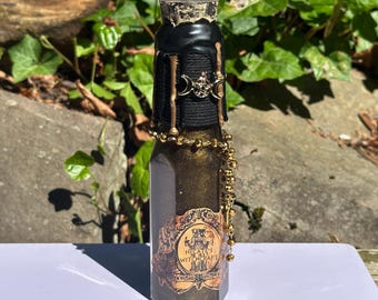 May include: A decorative glass bottle filled with a shimmering gold liquid. The bottle is adorned with a black leather-like wrap, gold accents, and a decorative label that reads "Hecate's Witchcraft". A gold beaded chain is draped around the bottle.