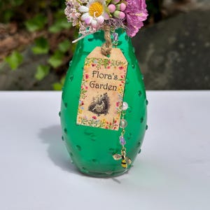 May include: A green glass vase with a textured surface, holding a bouquet of pink and white flowers. A tag with the text "Flora's Garden" and a bee charm are attached. The vase is approximately 15 cm tall.
