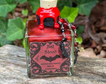 Puede incluir: Una pequeña botella de vidrio con un tapón de corcho rojo y un líquido rojo en su interior. La botella tiene una etiqueta que dice "Sangre de murciélago" y "Recolectada de murciélagos vampiros en Transilvania".