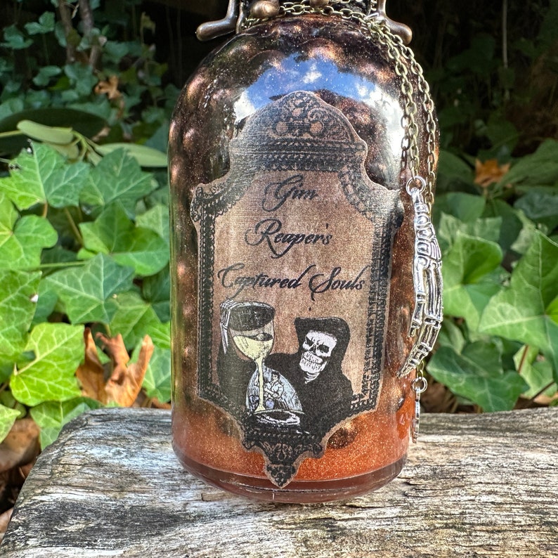 May include: A glass bottle with a gold glitter finish and a label that reads "Gin Reapers' Captured Souls". The label features a skeleton holding a goblet. A silver skeleton hand charm hangs from a chain around the bottle's neck.