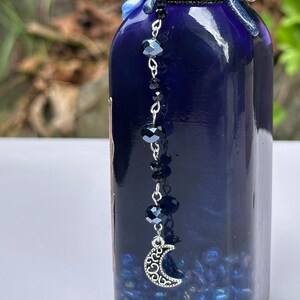 May include: A dark blue glass bottle with a decorative silver chain. The chain features black faceted beads and a crescent moon charm. The bottle is filled with small blue beads.