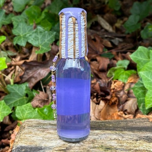 K&ouml;nnte beinhalten: Eine kleine Glasflasche mit einer violetten Fl&uuml;ssigkeit. Die Flasche ist mit einem wei&szlig;en und goldenen Wachssiegel versehen. Eine violette und goldene Perlenkette h&auml;ngt an der Flasche.