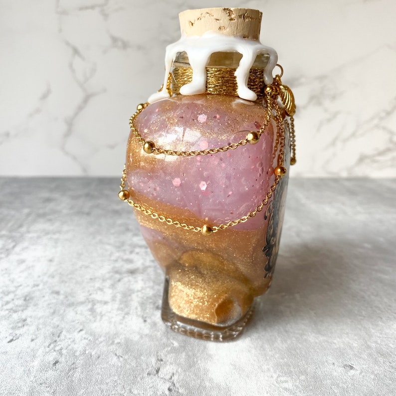 May include: A glass bottle with a pink and gold glitter mixture inside. The bottle is decorated with gold chains and beads, and a white wax seal on the cork.